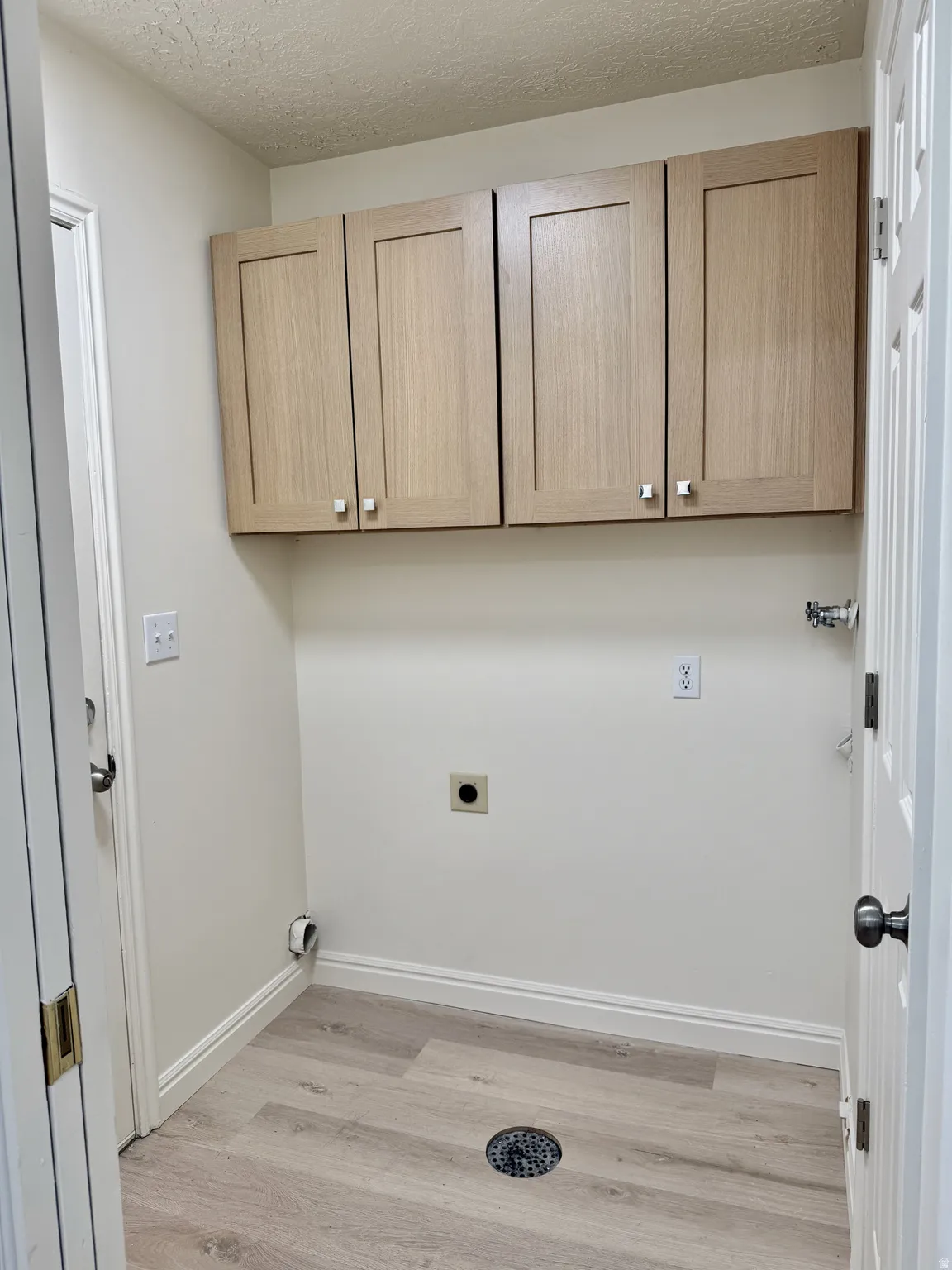 Dedicated utility space featuring light-toned wood-finish cabinetry, light wood-finish flooring, a floor drain, and white baseboards