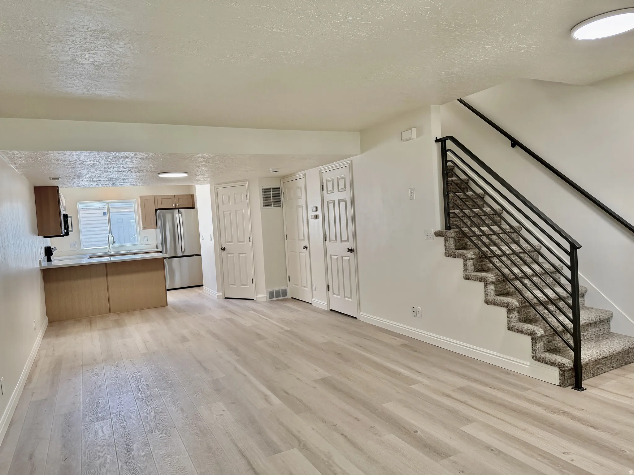 Open-concept living area with wood-finish flooring, a contemporary staircase featuring black metal railings and carpeted treads, and a kitchen area with stainless steel appliances, light-toned cabinetry, and a window with horizontal blinds