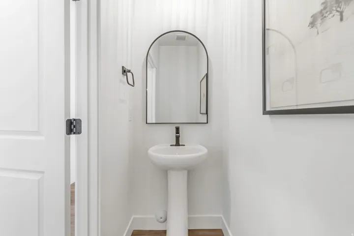 Pedestal sink with black wall-mounted faucet, arched mirror with black frame, and wood-finish flooring