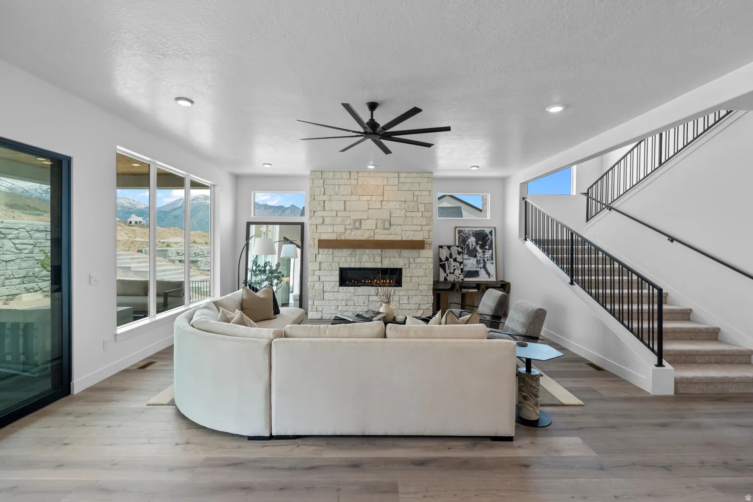 Spacious living area featuring a stone-clad fireplace with a wood mantel, a modern ceiling fan, and large windows showcasing mountain scenery