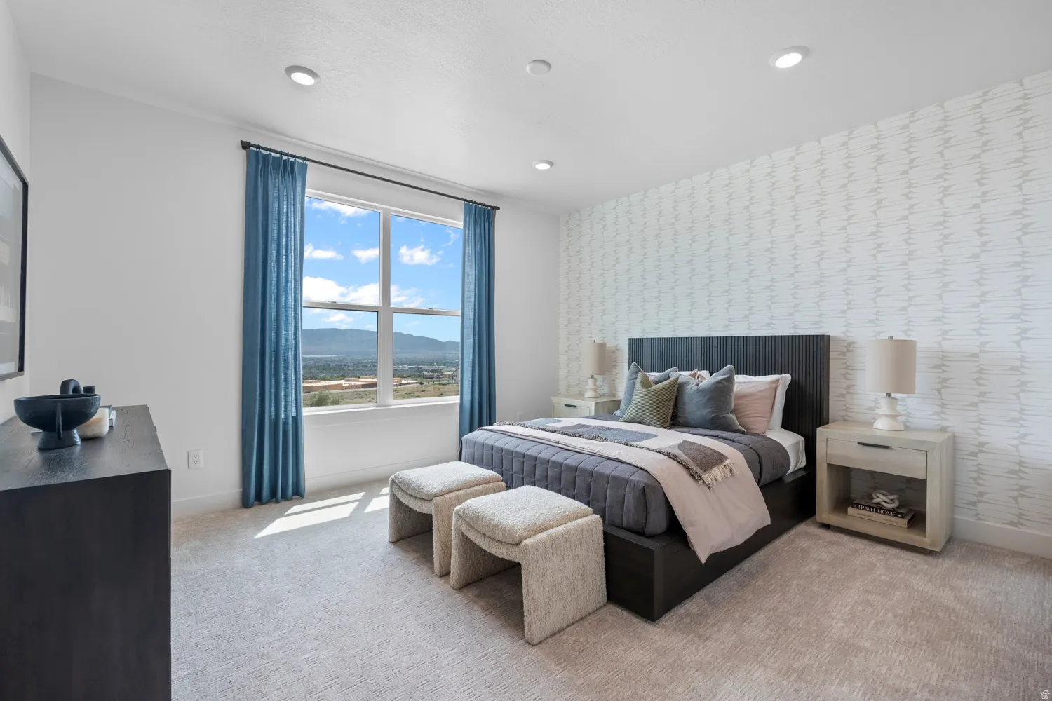 Expansive window offering a distant landscape backdrop, recessed ceiling lighting, textured accent wall, light-toned carpeting, and a dark wood-finish headboard