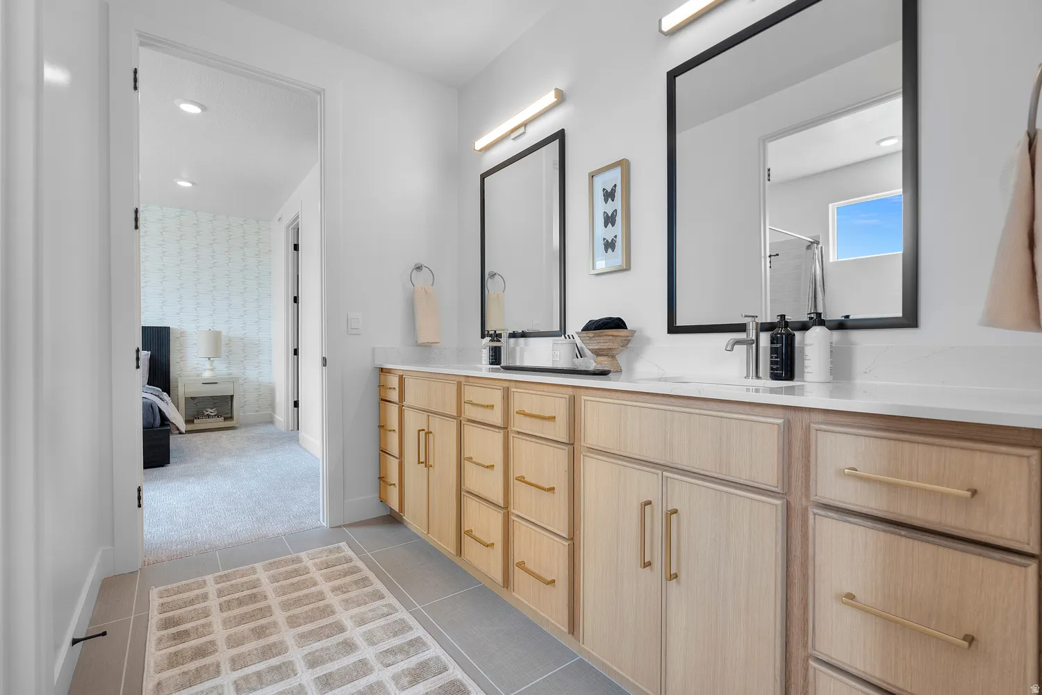 Double vanity featuring light wood-finish cabinetry with gold-tone hardware, white countertops, and dual rectangular mirrors with black frames