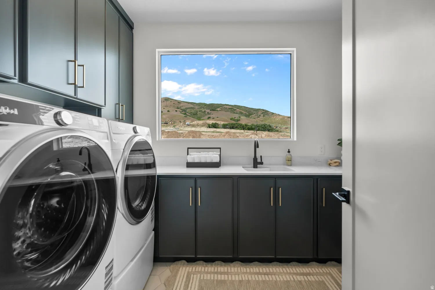 Modern laundry room featuring dark cabinetry with gold-tone hardware, a rectangular window, built-in sink with a matte black faucet, and light-toned countertops