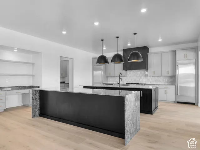 Kitchen featuring waterfall-edge stone countertops, custom cabinetry, three pendant light fixtures, a tile backsplash, and wood-finish flooring
