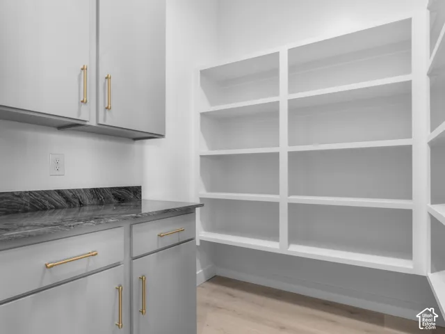 Walk-in storage space featuring built-in white shelving, gray cabinetry, contrasting dark stone-finish countertops, and metallic hardware