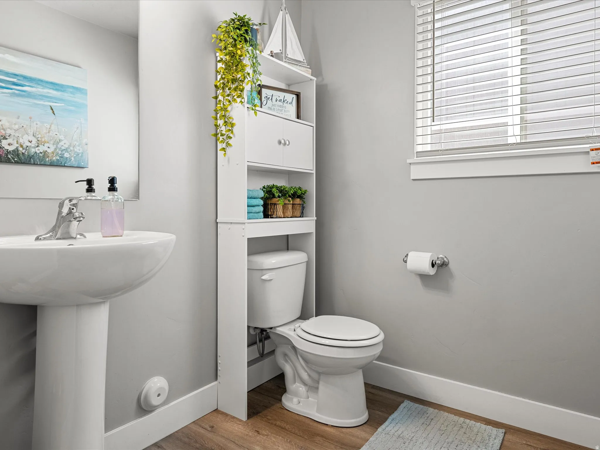 Pedestal sink with chrome faucet, wood-finish flooring, white baseboards, and a window with blinds