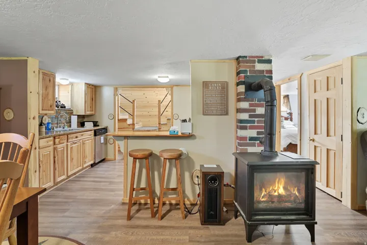 Wood-finish flooring throughout this space, featuring a wood-burning stove with a brick surround, a kitchen area with wood cabinetry, and a breakfast bar with wood stools