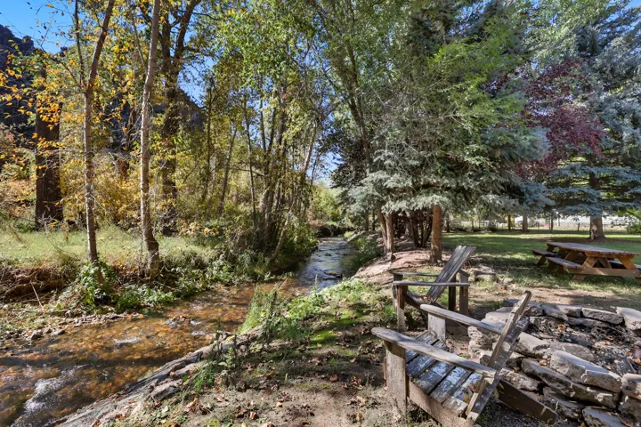 Stream-side setting featuring a natural rock fire pit, wooden Adirondack chairs, a picnic table, and mature trees