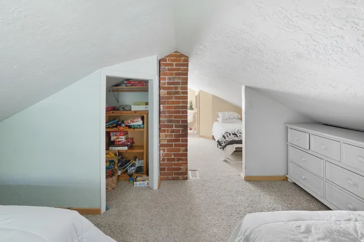 Carpeted loft area featuring a prominent exposed brick chimney, angled ceilings, and a built-in closet with wooden shelving