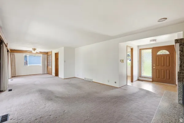 Spacious living area featuring light-colored carpeting, a ceiling fan, and a large window with white curtains