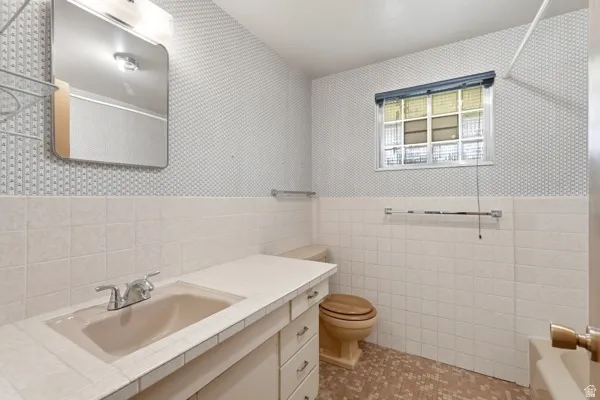 Bathroom featuring a vanity with an integrated sink, patterned wallpaper, a window with blinds, and tiled walls