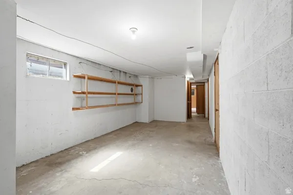 Unfinished basement featuring a concrete floor, painted cinder block walls, exposed ceiling joists, wood shelving, and a small window