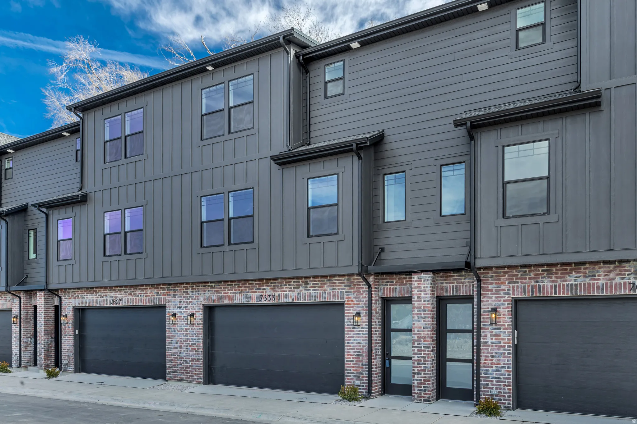 Modern townhome exterior featuring gray vertical siding, gray horizontal siding, brick accents, multiple windows with black frames, and a two-panel glass entry door