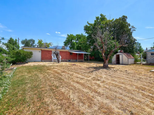 Expansive backyard featuring a brick exterior, white stairs, a covered patio, mature trees, and a storage shed