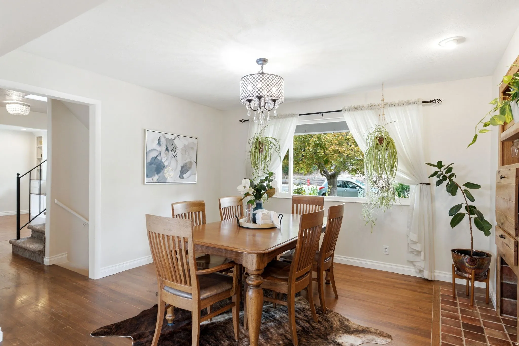 Dining area featuring wood-finish flooring, a contemporary chandelier, and a large window with white sheer curtains