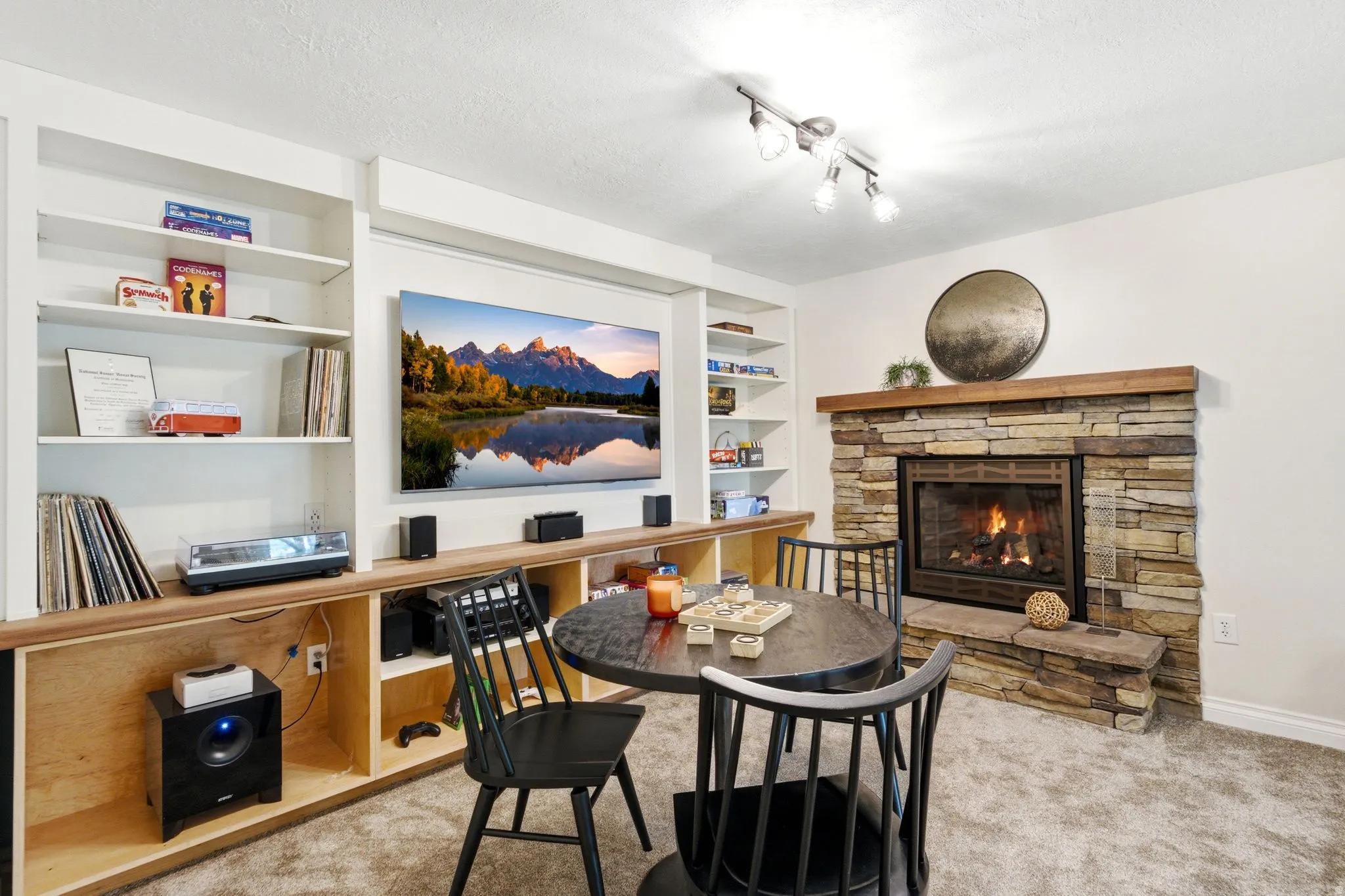 Living area featuring a stone-clad fireplace with a wood mantel, built-in shelving, track lighting, and soft carpet flooring