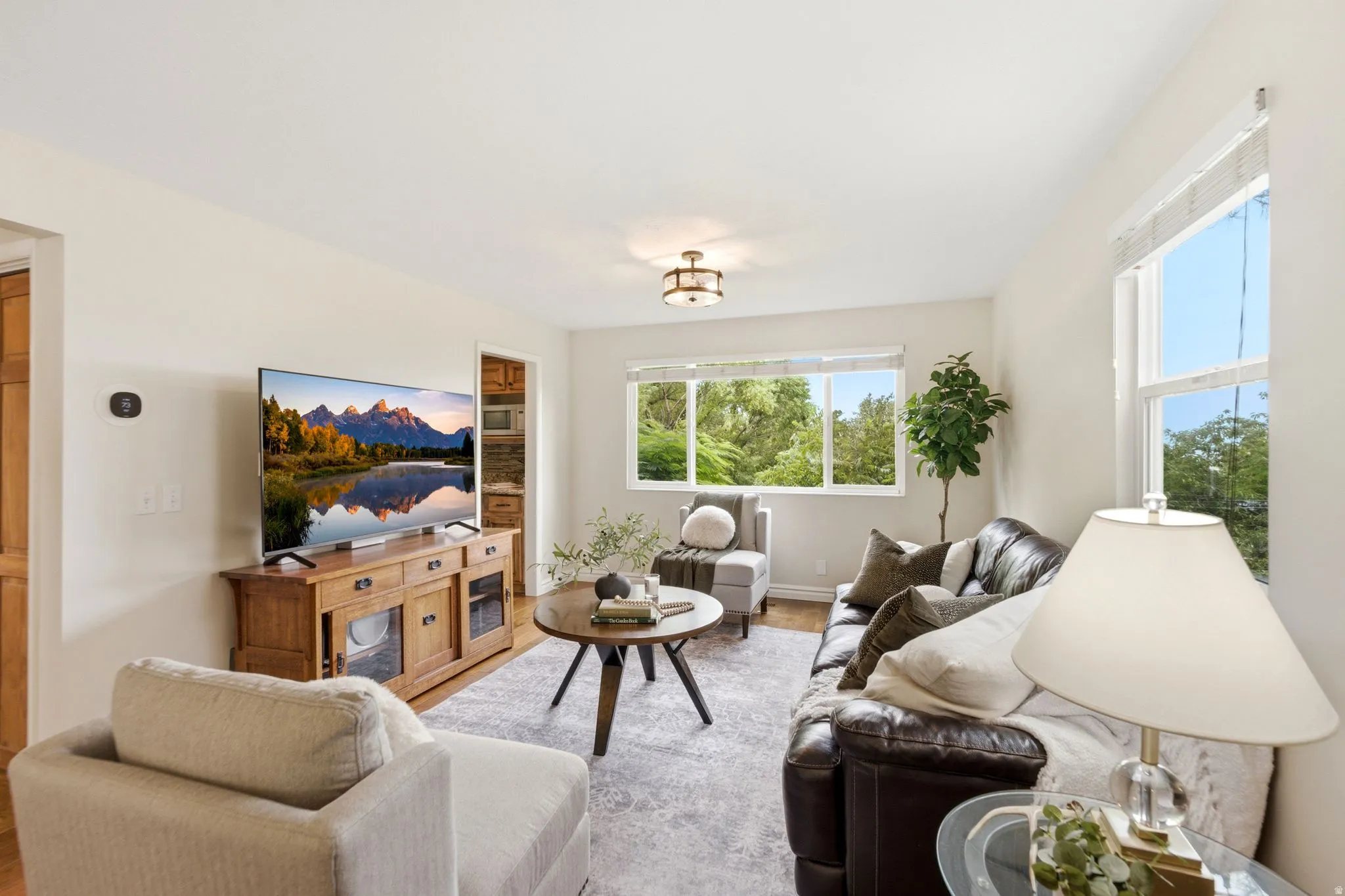 Bright living area with light-toned walls, wood-finish flooring, and multiple windows offering tree-lined scenery