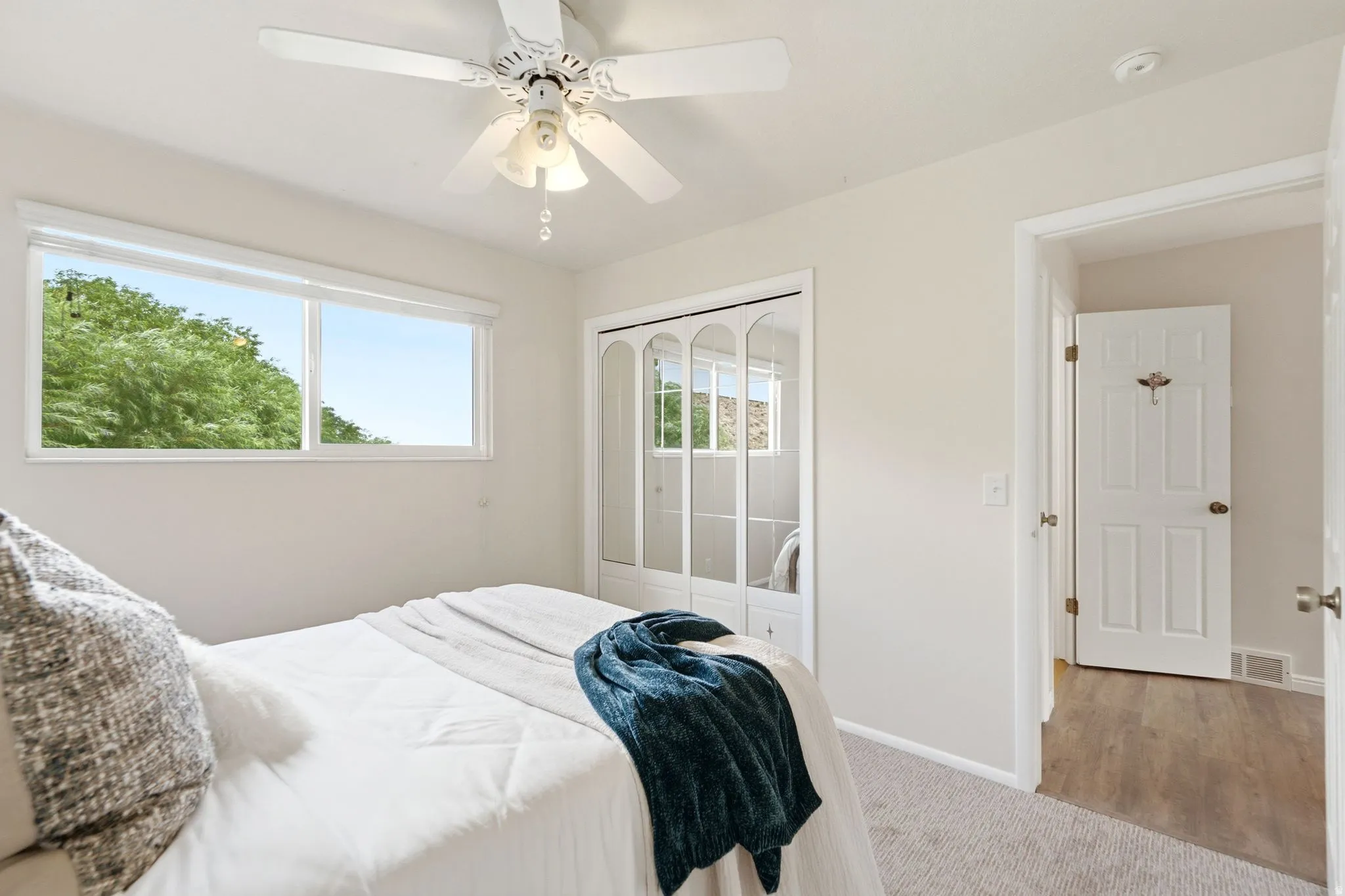 Bright room featuring a window with green tree scenery, a ceiling fan, mirrored closet doors, and light neutral wall paint