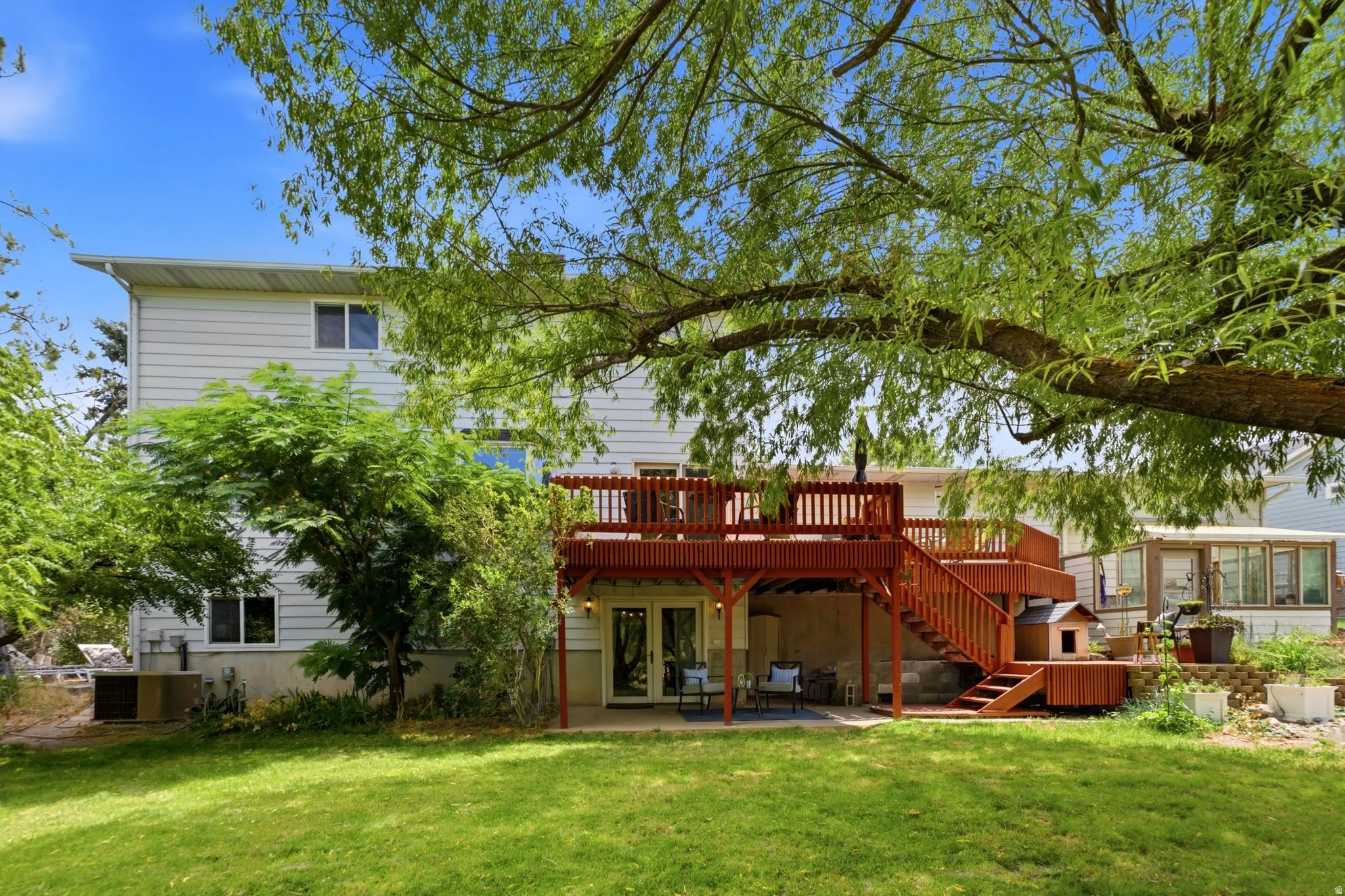 Spacious backyard featuring a multi-level wooden deck with balustrade railings and stairs, a lower patio area with double glass doors, mature trees providing natural shade, and a vibrant green lawn