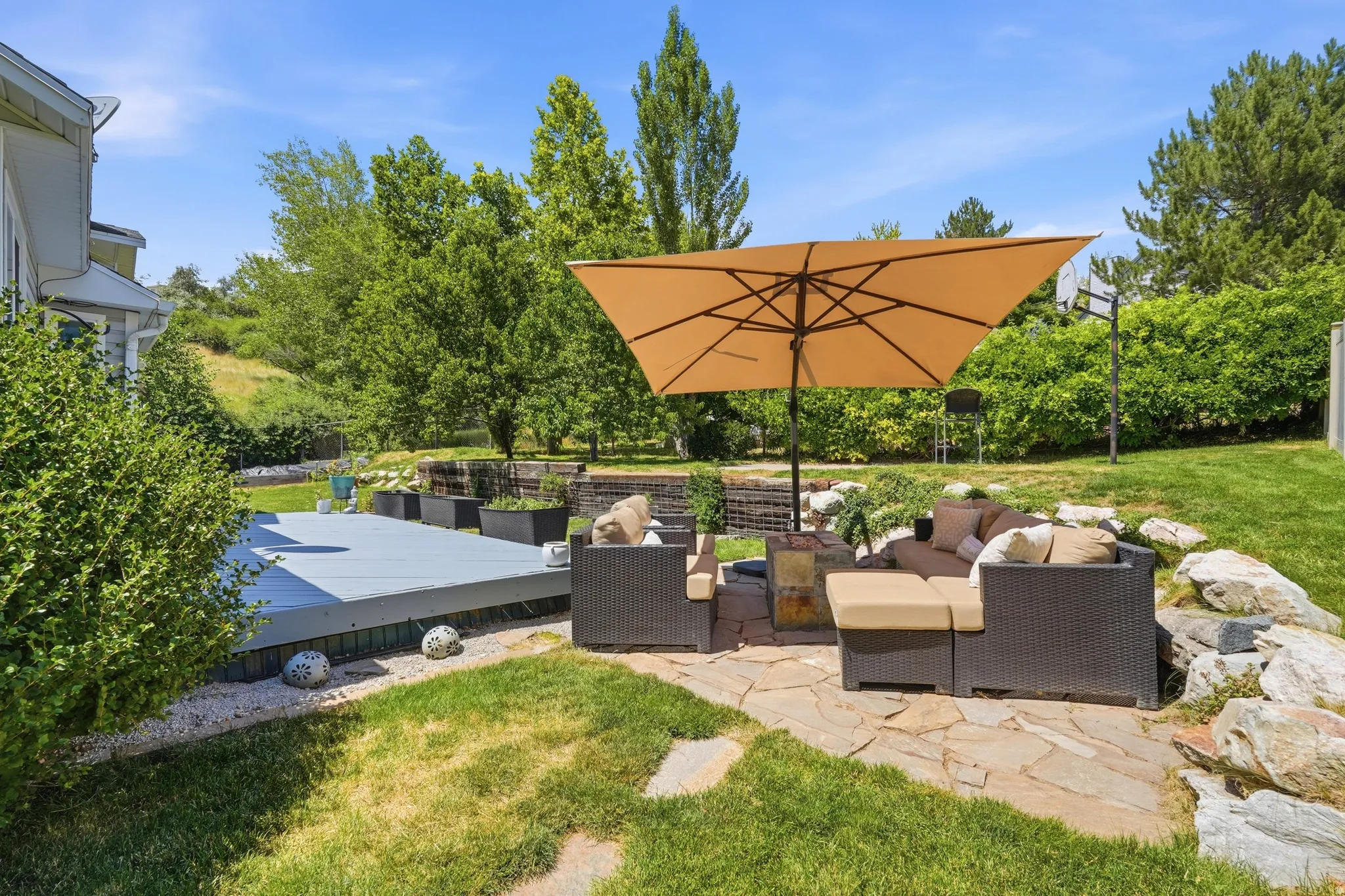 Expansive outdoor area featuring a grey-toned deck, a flagstone patio, mature landscaping, and a sloped, tree-lined background