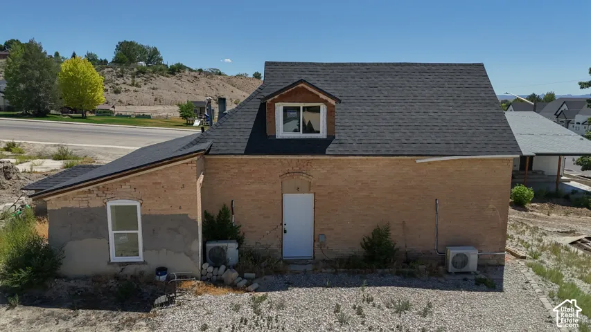 Brick exterior structure featuring a dormer window, dark shingle roofing, a white entry door, and multiple air conditioning units