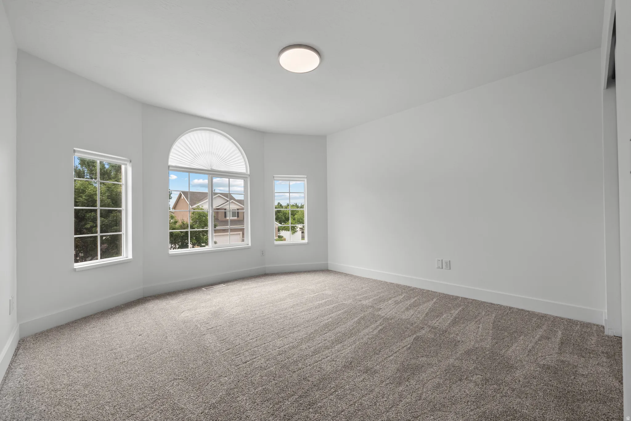 Bright room with gray carpet flooring and white walls, featuring a bay window configuration with a prominent arched window, two rectangular windows, and a flush-mount ceiling light fixture