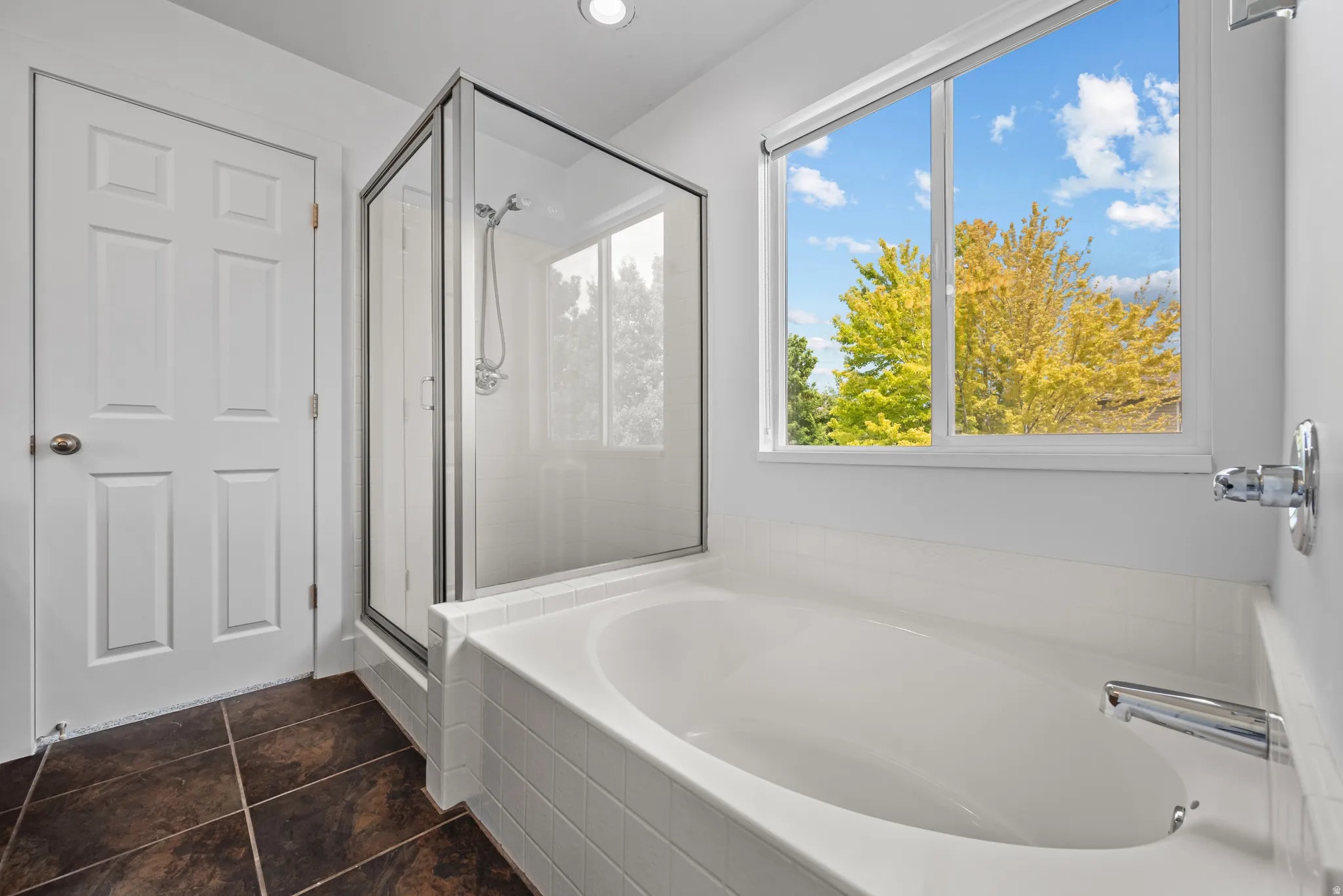Soaking tub with white tile surround, glass-enclosed shower, and a large window