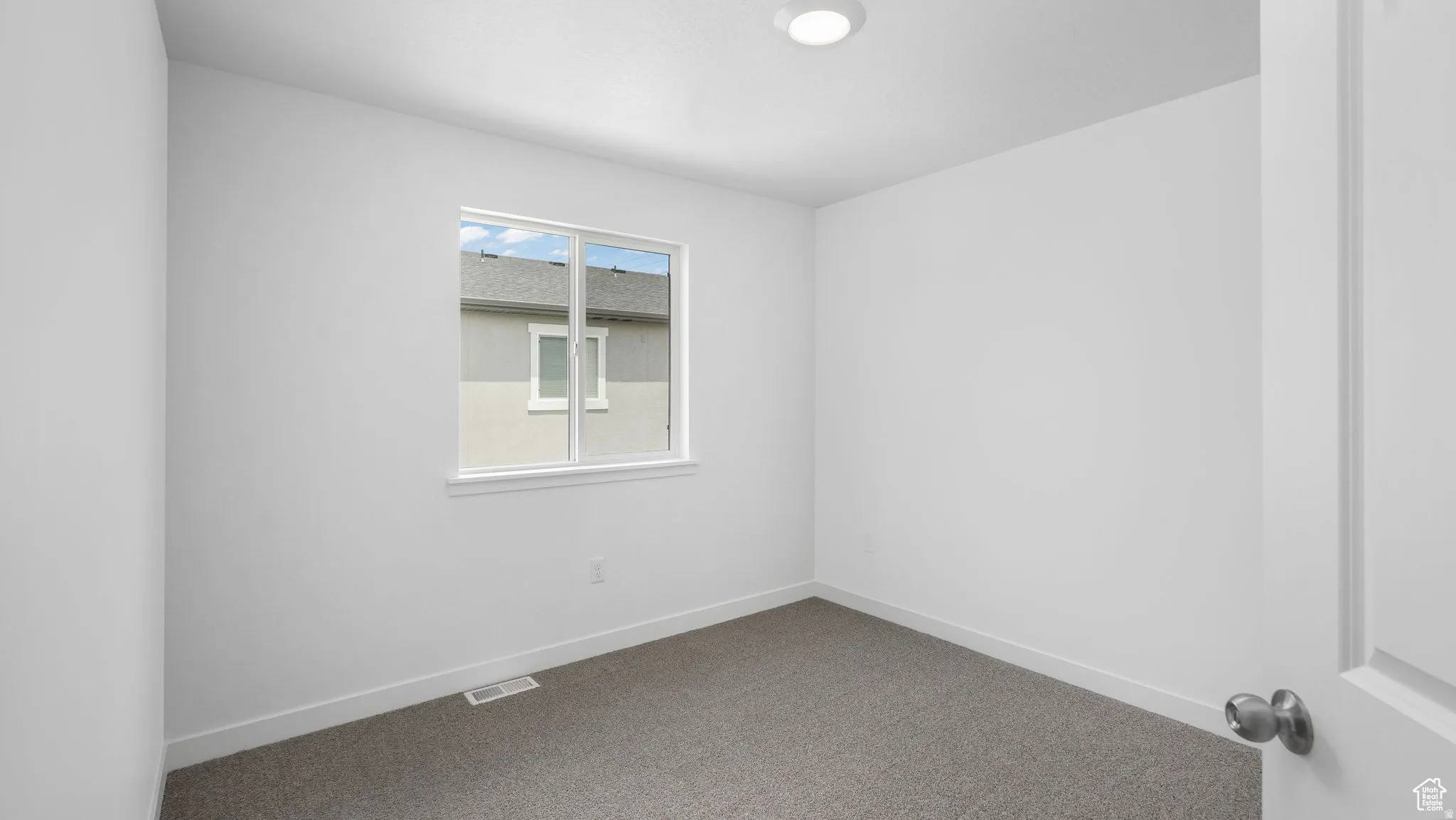 Neutral white walls throughout, showcasing a window with white trim, gray carpeting, and a recessed ceiling light fixture