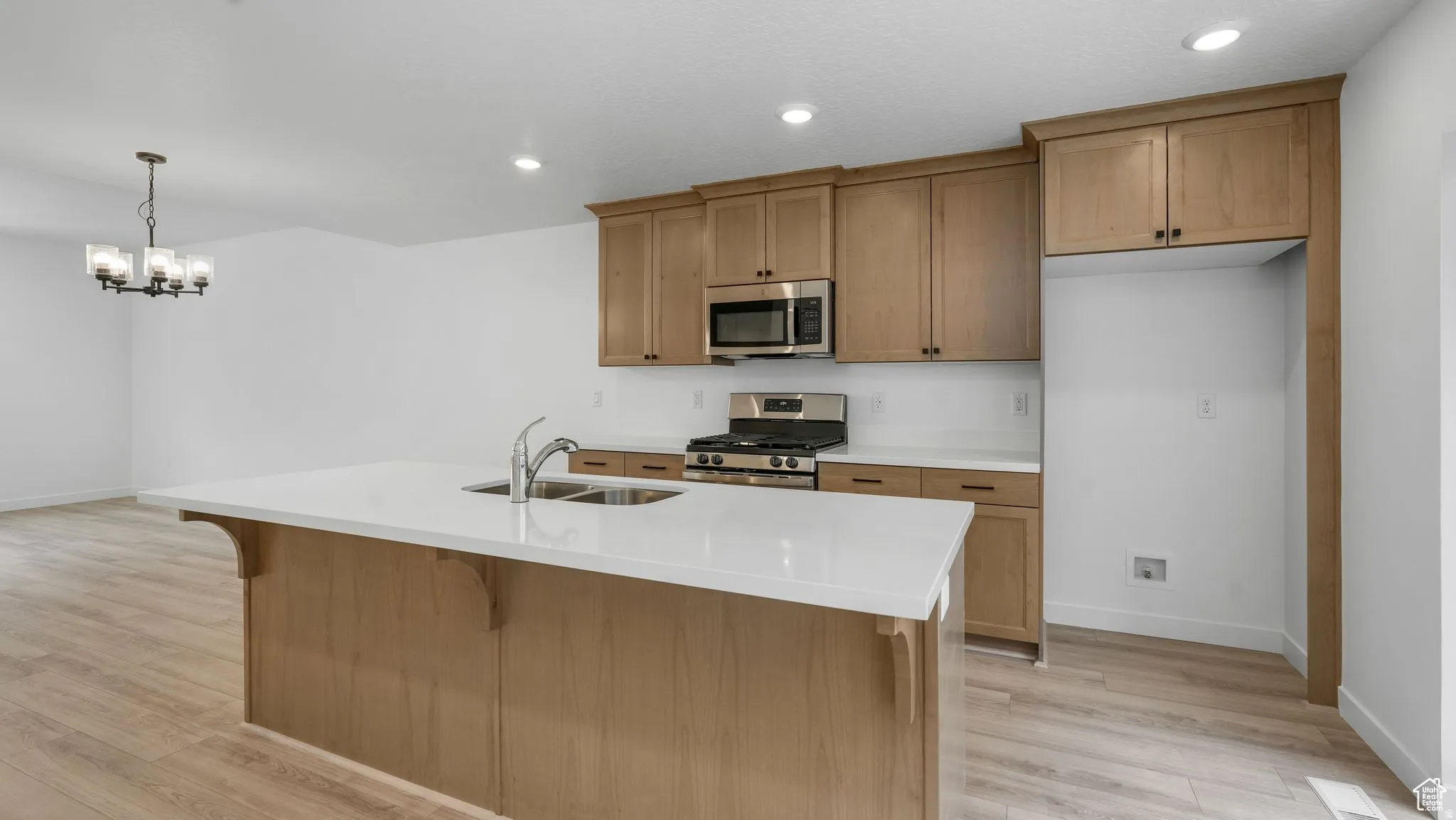 Contemporary kitchen featuring a large central island with a double basin sink, light wood-finish cabinetry, stainless steel appliances, and wood-finish flooring