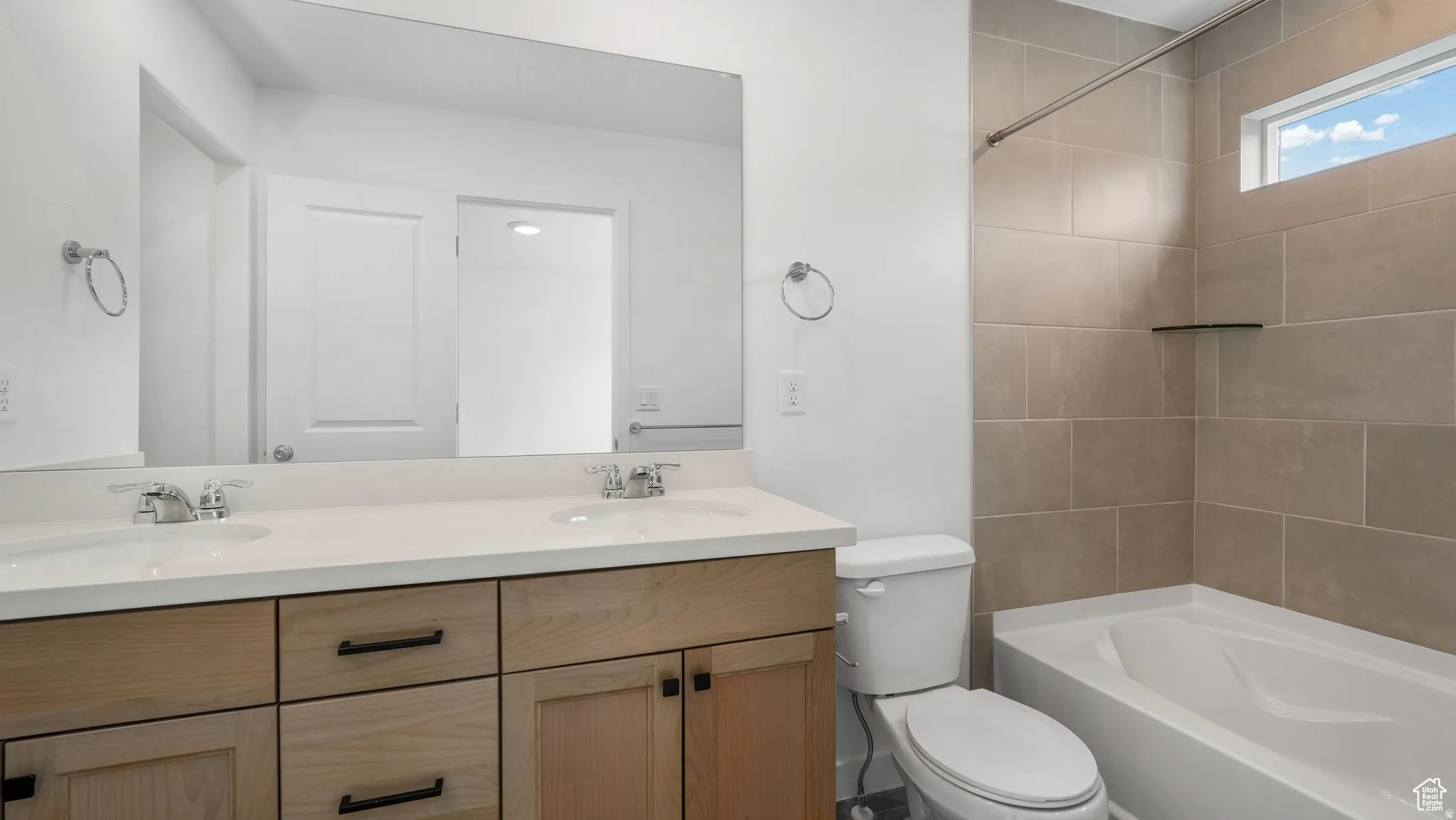 Modern bathroom featuring a dual vanity with light wood-finish cabinetry, a large mirror, a tiled shower-over-tub combination, and a horizontal window