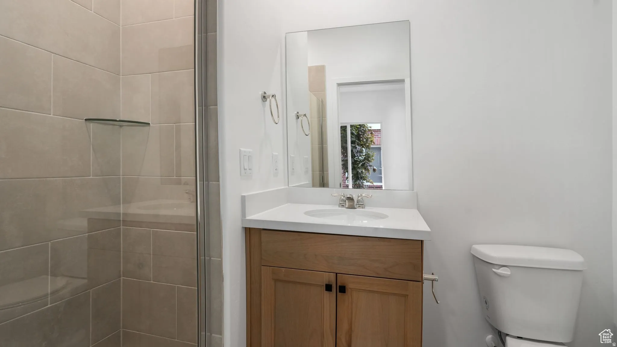 Bathroom featuring a glass-enclosed shower, light-toned tile, a floating corner shelf, and a vanity with a white countertop