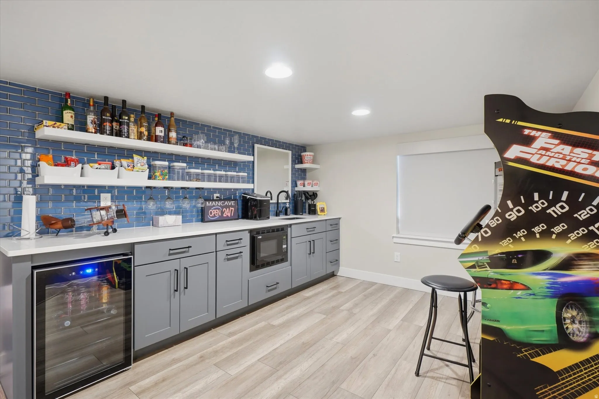 Wet bar featuring a blue subway tile backsplash, white floating shelves, light gray cabinetry with dark hardware, and a built-in beverage refrigerator