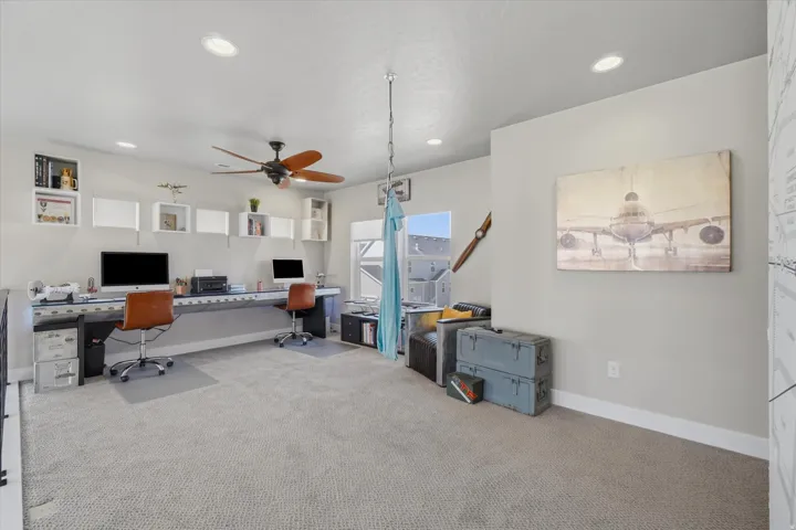 Carpeted loft space featuring a built-in desk with dual workstations, white wall-mounted shelving, and a ceiling fan with integrated lighting