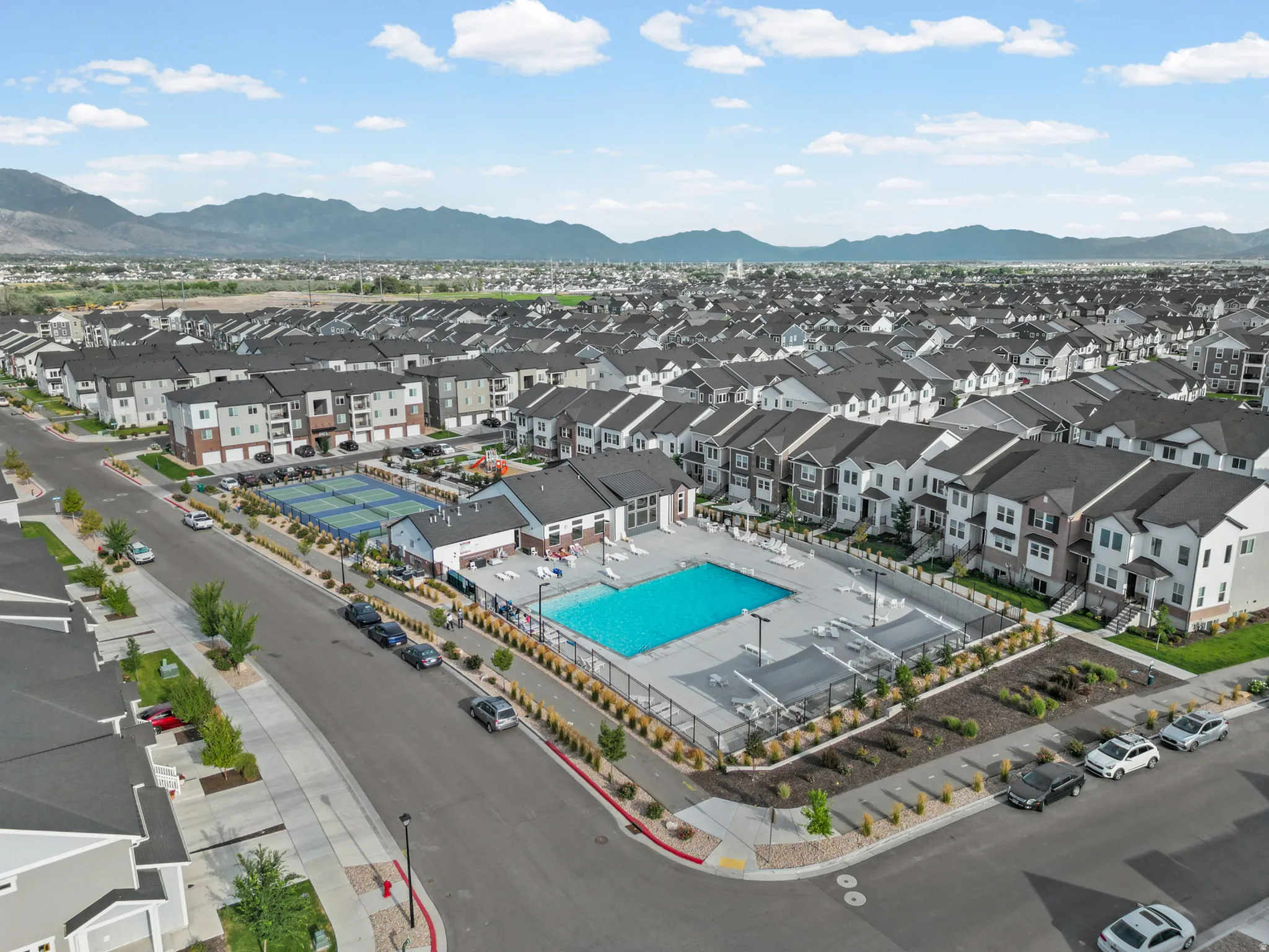 Aerial perspective showcasing a residential community with a large outdoor swimming pool, adjacent recreational building, and two tennis courts