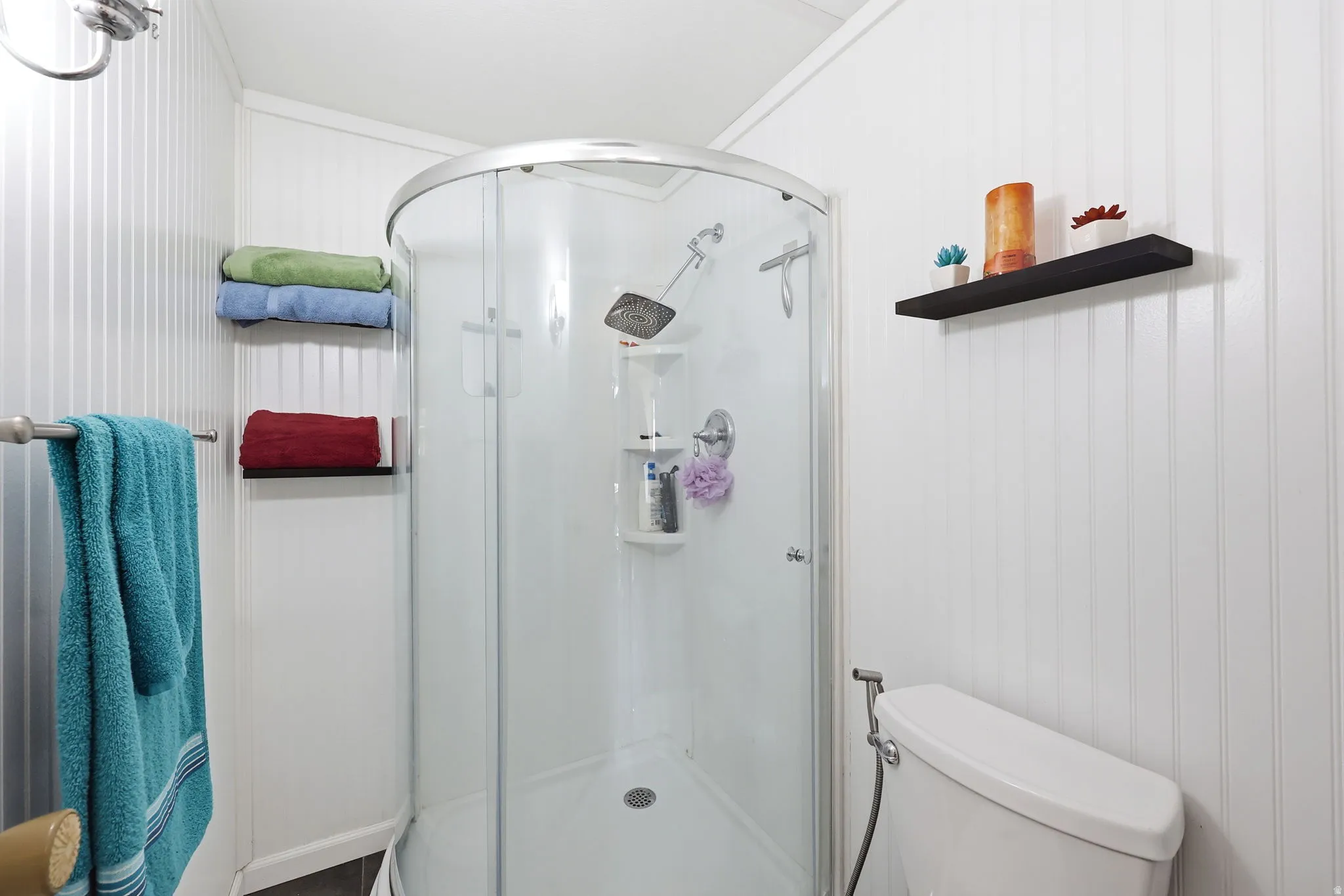 Bathroom featuring a rounded glass-enclosed shower with a rainfall showerhead, white beadboard-style walls, and dark wood-finish flooring