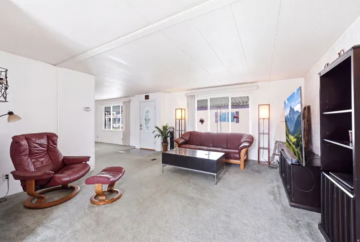 Spacious living area featuring light-colored wall panels, textured ceiling, and light gray carpeted flooring