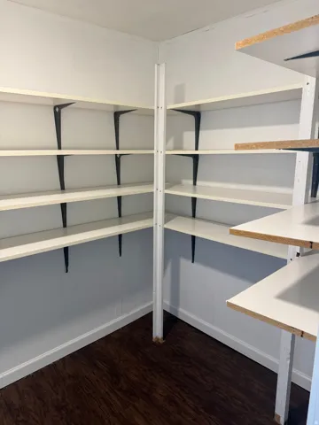 Corner pantry featuring multi-level white shelving, dark wood-finish flooring, white baseboards, and light-toned walls
