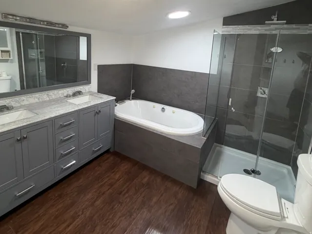 Bathroom featuring a dual vanity with a light-toned countertop, a built-in jetted tub, a glass-enclosed shower, and wood-finish flooring