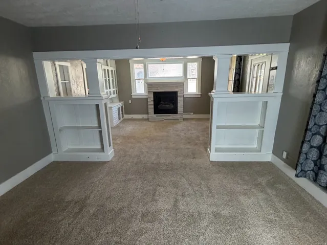 Spacious carpeted room featuring a ceiling fan with integrated lighting, built-in shelving, and an arched entryway leading to an additional space with a fireplace