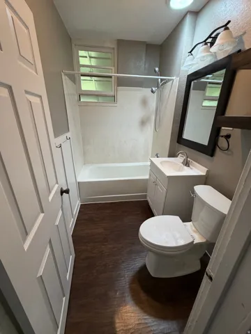 Bathroom featuring wood-finish flooring, white paneled door, single vanity with integrated sink, white toilet, and a white bathtub with shower