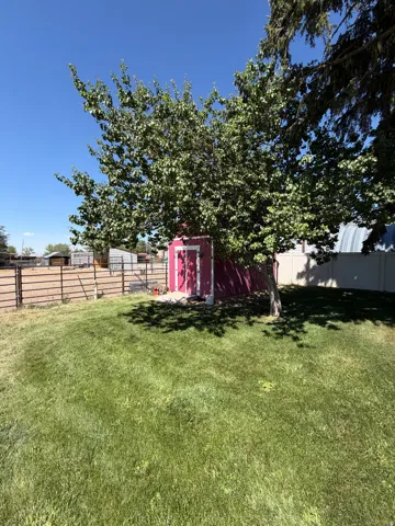 Expansive grassy yard featuring a red storage shed with a white door, mature deciduous trees, and a perimeter fence
