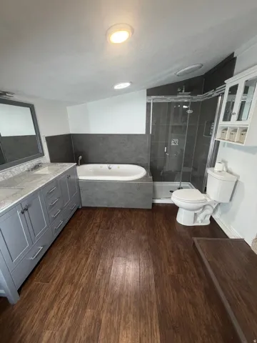 Bathroom featuring dark wood-finish flooring, a dual vanity with light gray cabinetry and light-toned countertops, an oval soaking tub, and a glass-enclosed shower with dark gray tile