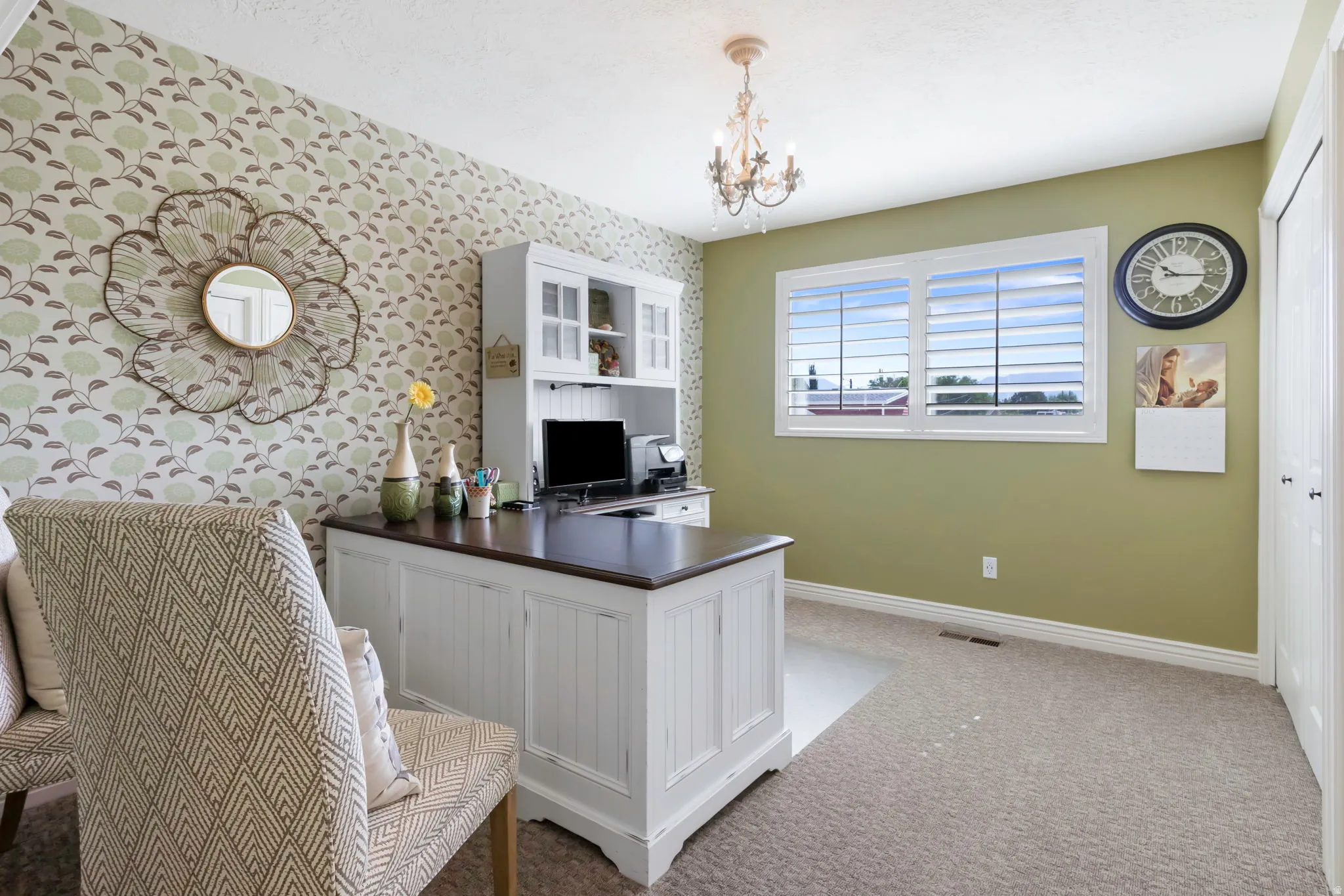 Dedicated workspace featuring a built-in white desk with a dark countertop and hutch, patterned accent wall, light green wall finishes, and a chandelier light fixture