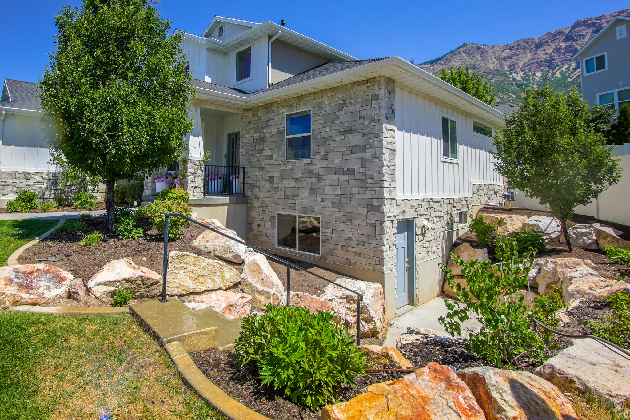 Contemporary residence featuring a stone and vertical siding exterior, a front porch with black railing, terraced landscaping with mature trees and large boulders, a concrete walkway with handrails, and a lower-level access door