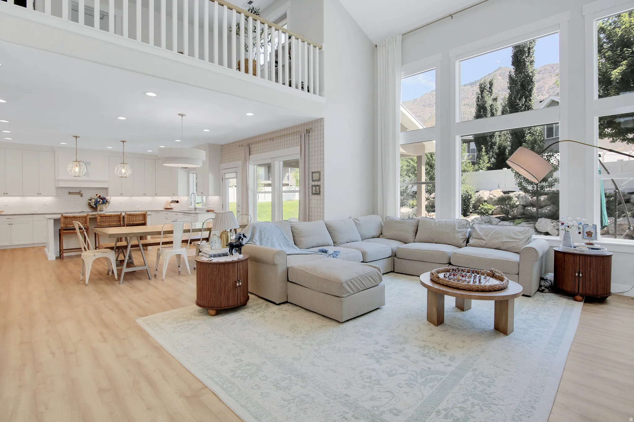 Expansive living area with high ceilings, featuring a two-story window array with mountain scenery, wood-finish flooring, and a kitchen with white cabinetry and pendant lighting