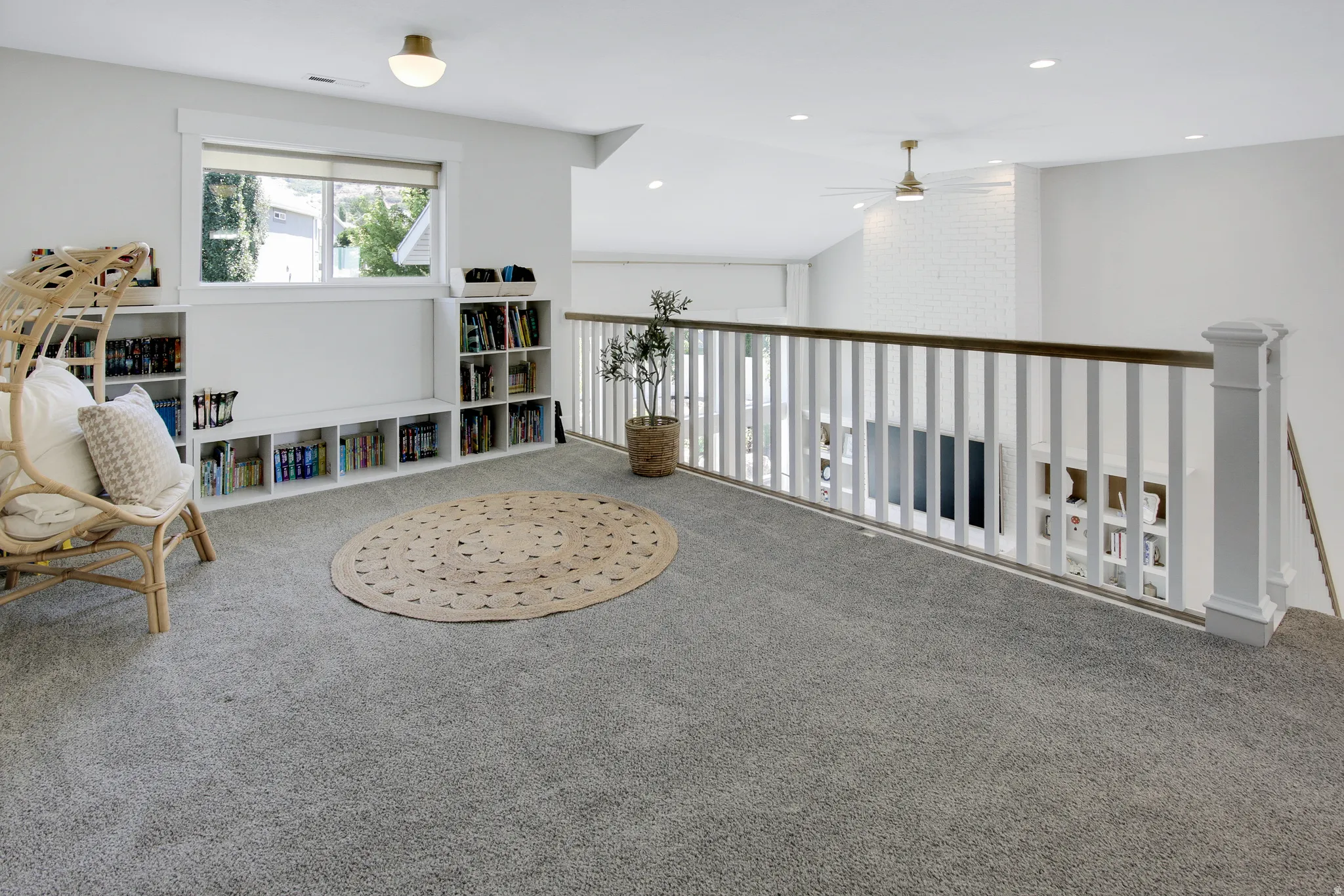 Carpeted loft area featuring a window with a white frame, built-in shelving, a white spindle railing with a wood-finish handrail, and an overhead light fixture