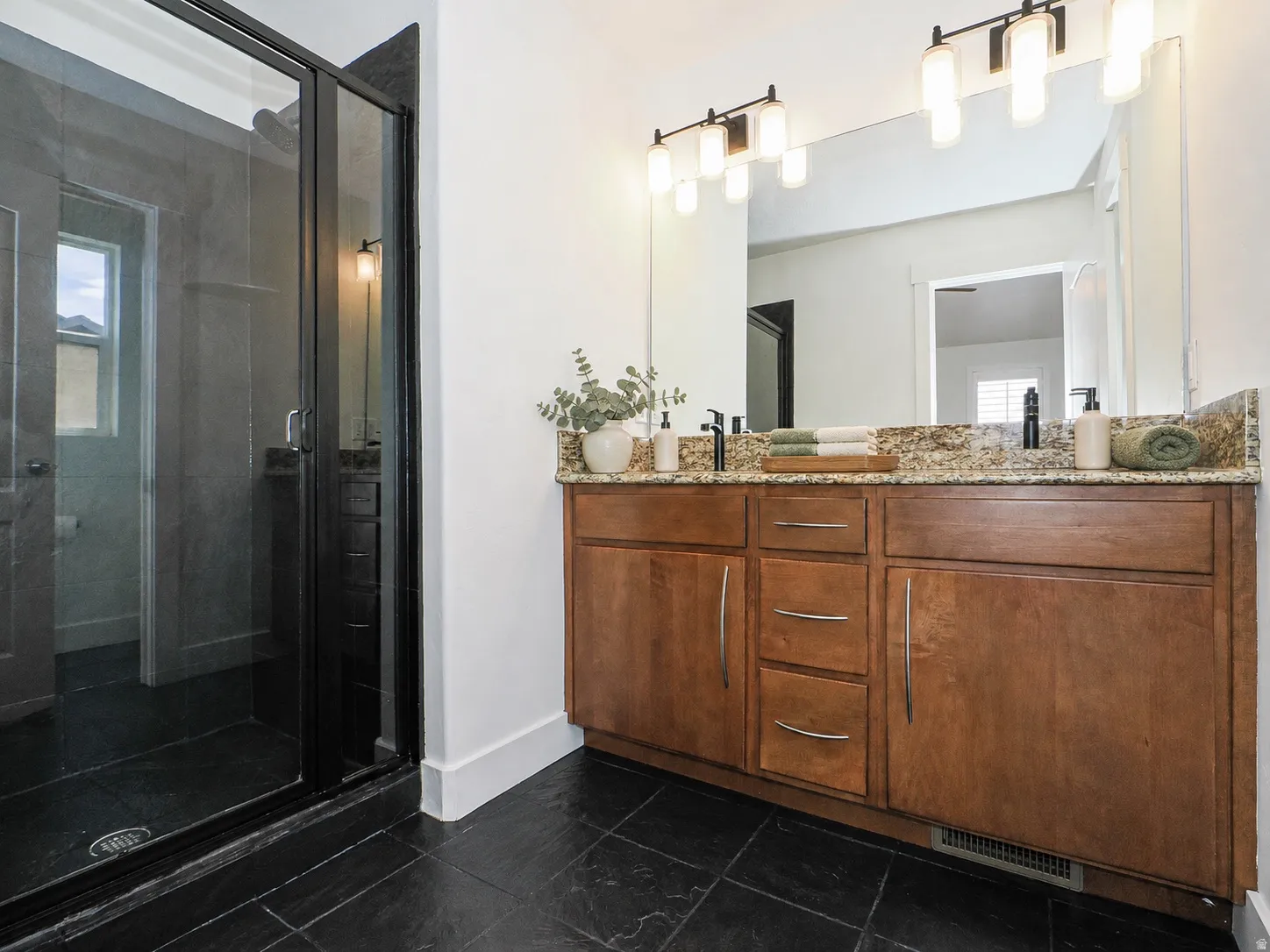 Walk-in shower with black-framed glass enclosure, dual sink vanity with stone countertop, wood cabinetry, and black tile flooring