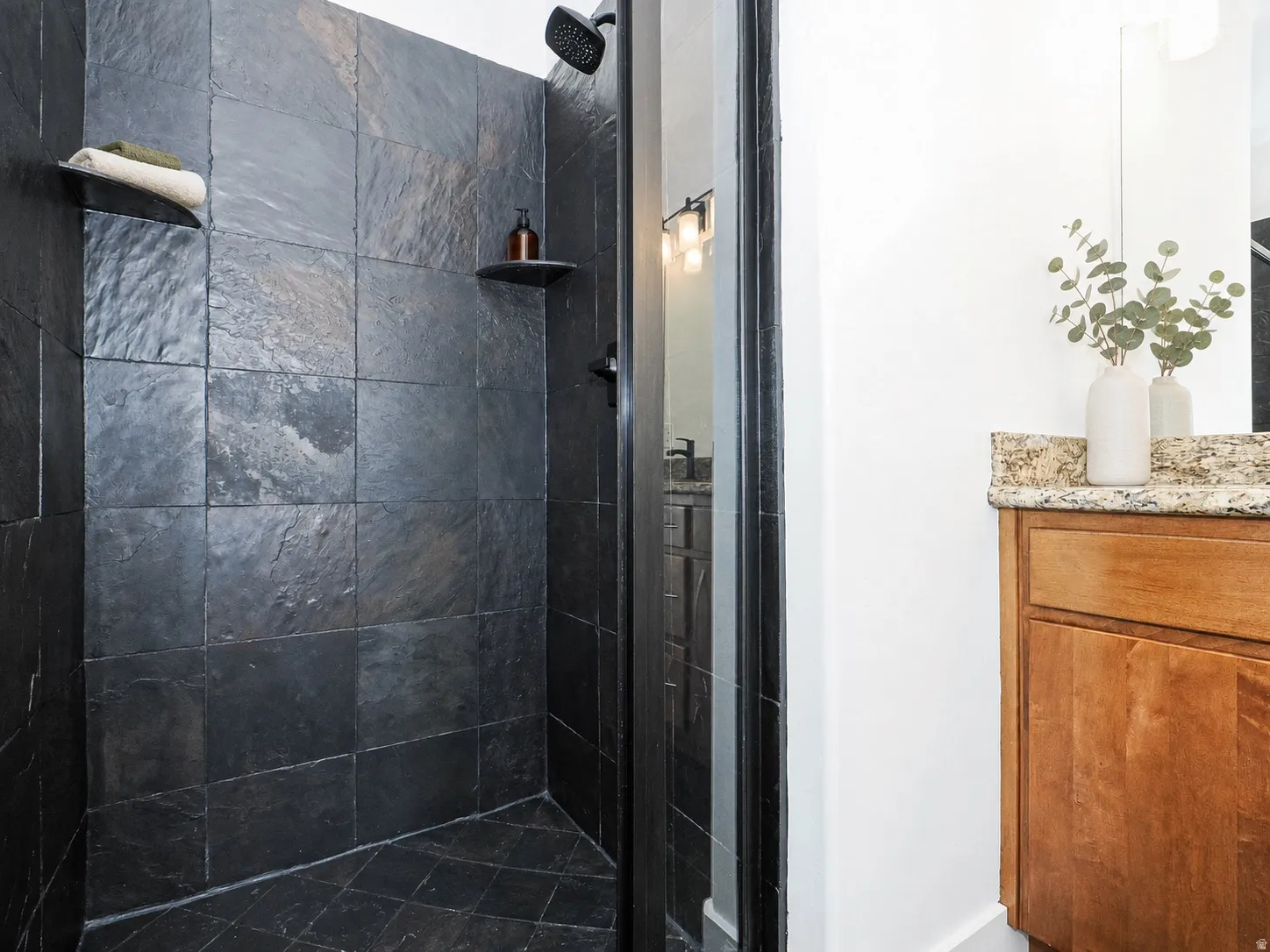Shower enclosure with dark stone-finish tile walls and floor, featuring a black shower head and integrated shelving