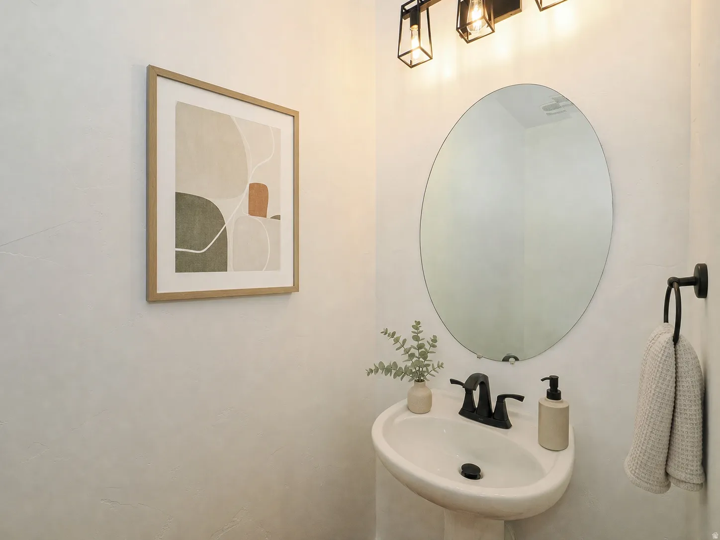 Pedestal sink featuring a matte black faucet, oval mirror, and three-light vanity fixture