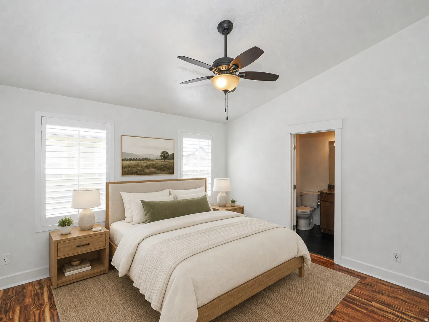 Bedroom featuring wood-finish flooring, two large windows with plantation shutters, a ceiling fan with integrated lighting, and a doorway leading to a bathroom with a toilet and vanity